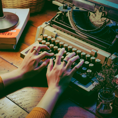 Hands typing on an old-fashioned typewriter, surrounded by books and a small plant on a wooden desk, creating a nostalgic, vintage writing atmosphere. Website Design and Copy