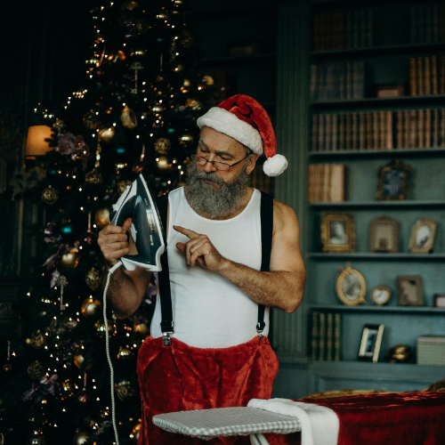 Man dressed as Santa Claus in suspenders and Christmas pants, ironing clothes in front of a beautifully decorated Christmas tree, with bookshelves in the background.