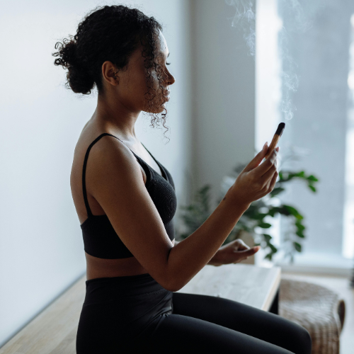 A woman with curly hair in a low bun, wearing a black sports bra and leggings, is sitting cross-legged on a wooden surface, holding a lit incense stick. She is focused and appears to be meditating or practicing mindfulness in a serene, minimalist room with natural light filtering in. Shes getting ready to hire House of Bettencourt for her Spiritual Website Design