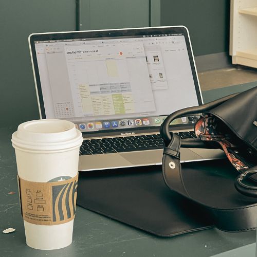 A laptop displaying a calendar with a cup of coffee and a bag placed next to it on a table. The workspace suggests a productive setting, ideal for planning or organizing tasks for real estate website development.