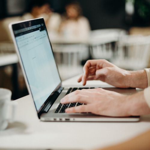 A close-up of a person's hands typing on a laptop at a table in a cozy café. The background is softly blurred, allowing the focus to remain on the individual working on the computer.