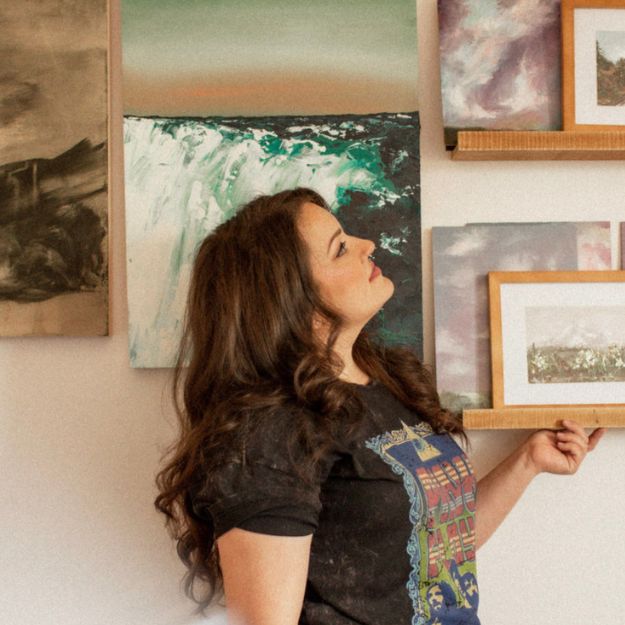 An artist stands in front of their displayed artwork, gazing thoughtfully at a wall filled with framed and unframed paintings. The display features abstract and landscape pieces, creating a visually artistic setup.