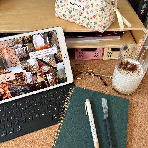 A cozy workspace featuring a tablet displaying an autumn-themed home screen, a notebook, pens, a glass of iced coffee, and a floral pouch. The setup creates a warm and inviting atmosphere, perfect for brainstorming or planning marketing strategies.
