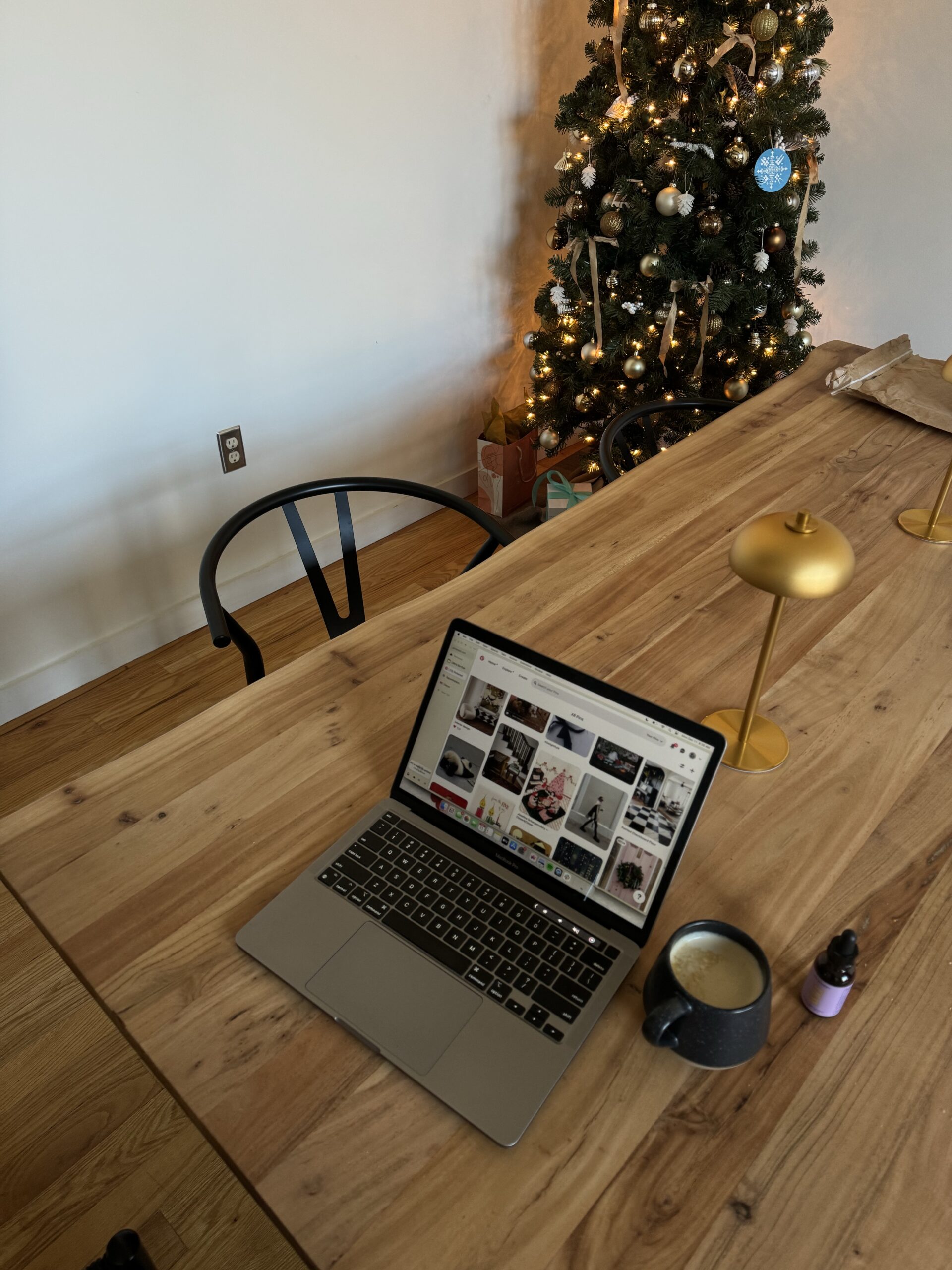 A cozy workspace with a laptop displaying a Pinterest board, a mug of coffee, a small bottle, and a decorated Christmas tree in the background.