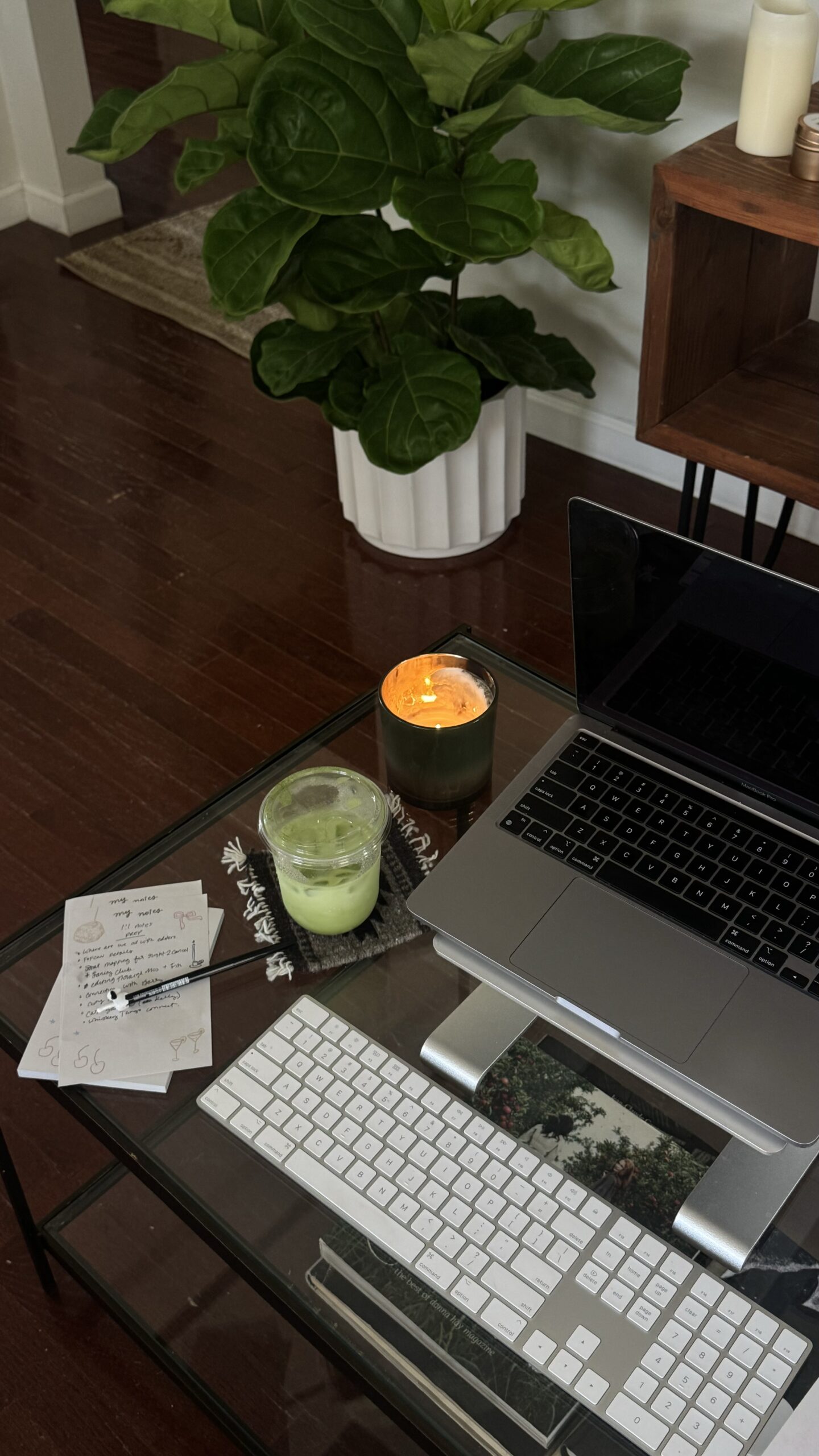 A cozy workspace featuring a laptop, external keyboard, a notepad with handwritten notes, a green iced matcha drink, and a lit candle on a glass coffee table. A fiddle-leaf fig plant in a white pot sits in the background, adding a touch of greenery to the modern, minimal aesthetic.