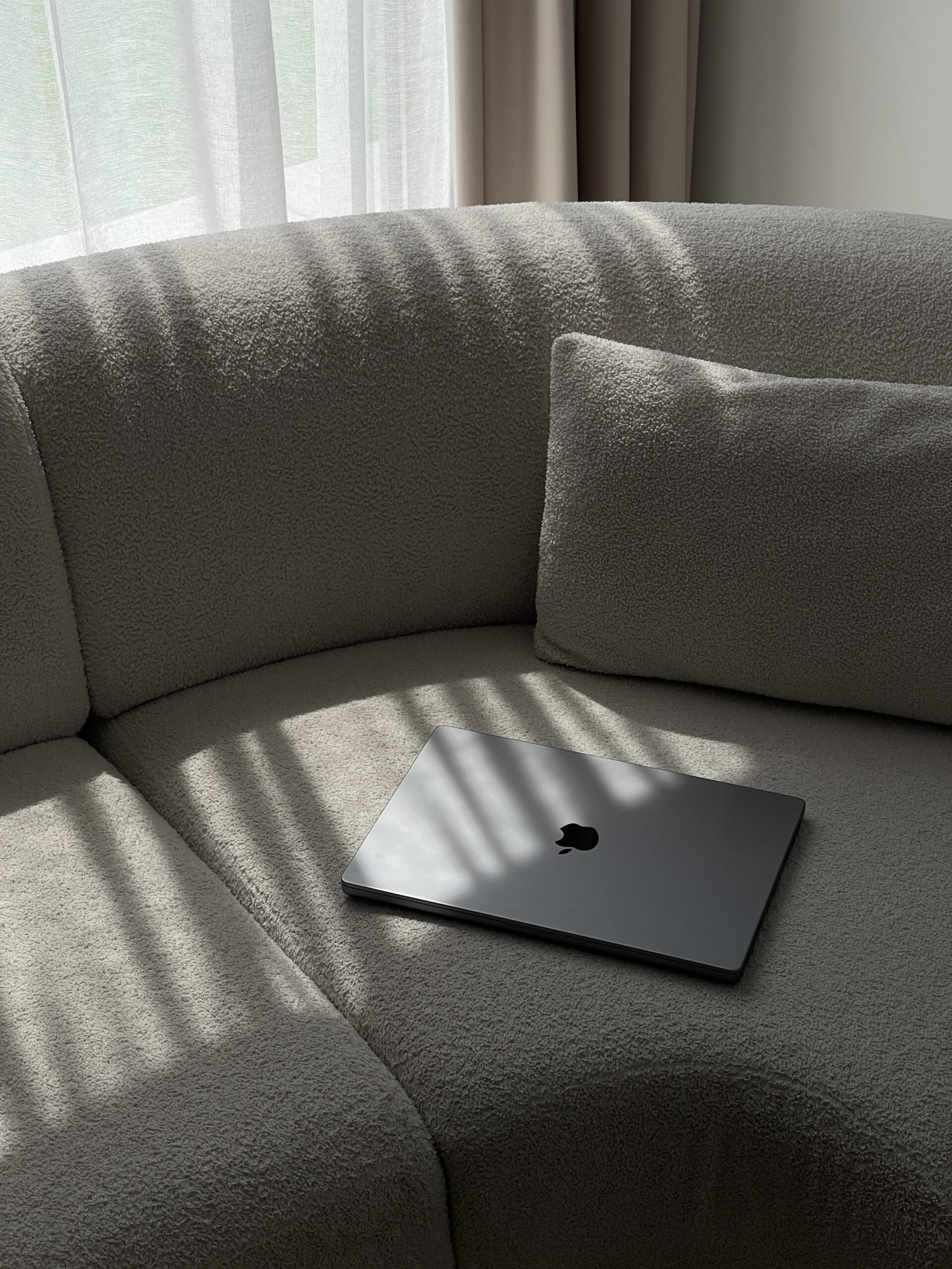 A closed MacBook rests on a modern, curved beige sofa with soft sunlight streaming through sheer curtains, casting elegant shadows. The scene exudes calm and creativity, ideal for a designer’s workspace.