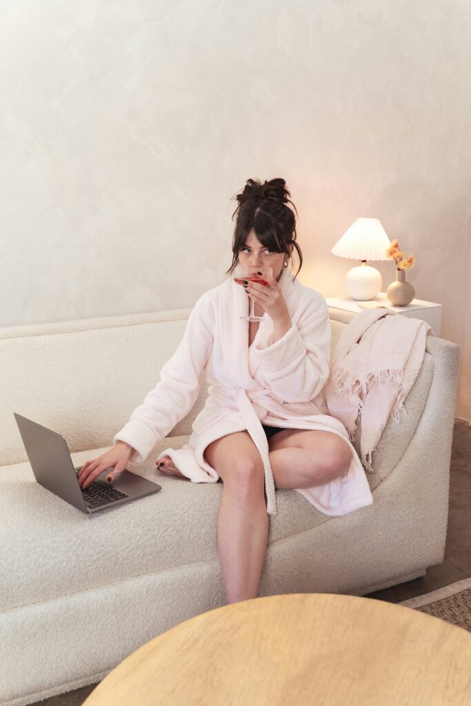 Brunette woman in a pink robe sitting on a cream sofa, using a laptop and sipping a drink, representing relaxed blogging workflow while considering how often should I blog for SEO