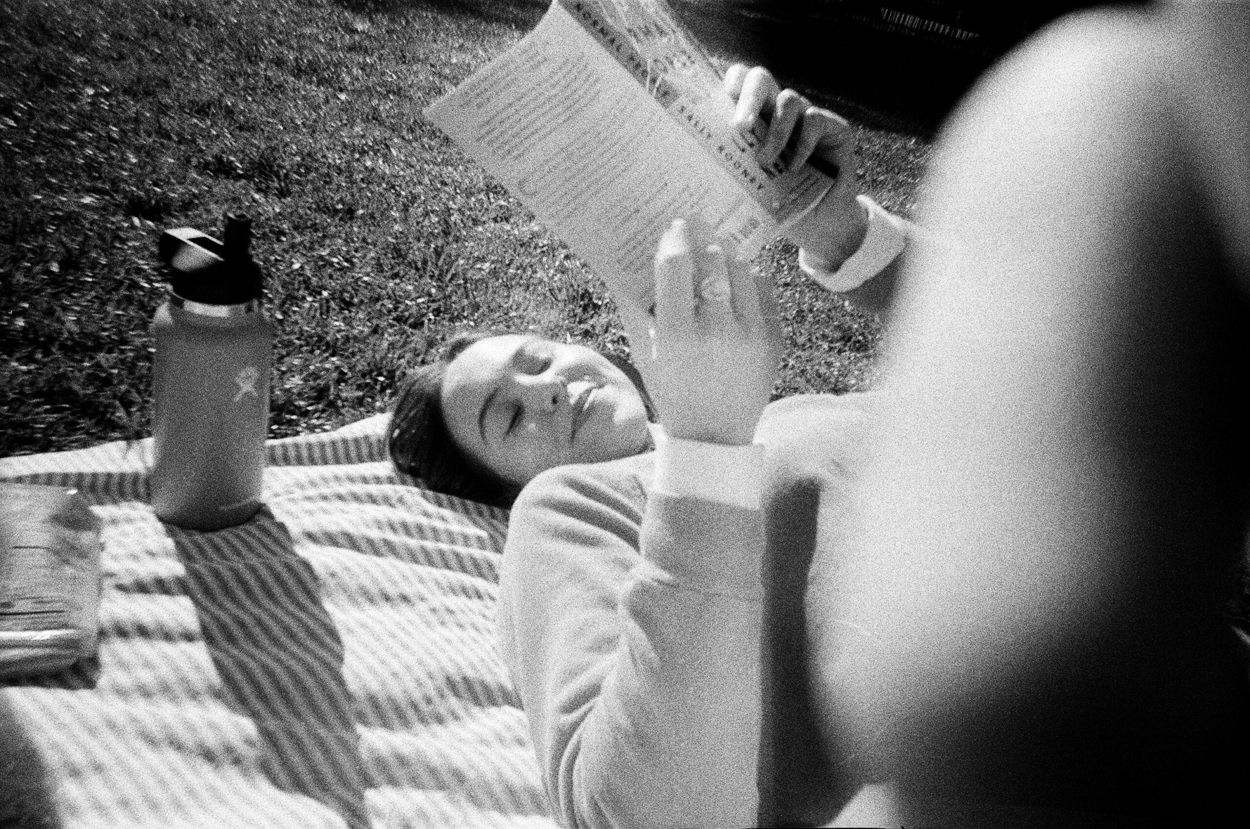 Black-and-white candid photo of a person lying on a picnic blanket outdoors, smiling while reading a book, with a water bottle beside them—relaxed lifestyle image that feels authentic and human.